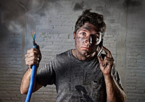 young man holding electrical cable smoking after electrical accident with dirty burnt face in funny desperate expression calling with mobile phone asking for help in electricity DIY repairs danger concept